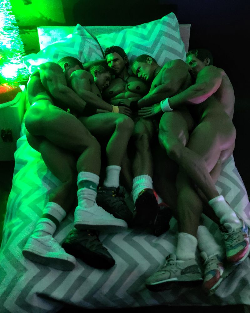 5 semi-naked men, lying on a bed, embracing each other. The men are muscular and wear sneakers and white socks. A Christmas tree bathes them in a green glow.
The image has been created using dolls.
