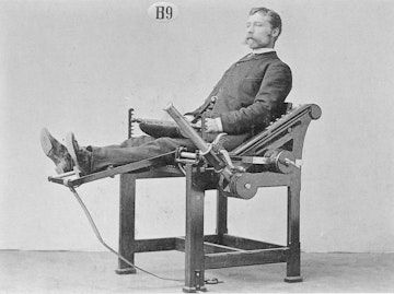 A moustachioed man in Victorian era suit and shoes sits in a incredibly primitive piece of exercise equipment with his legs raised as though in a recliner.