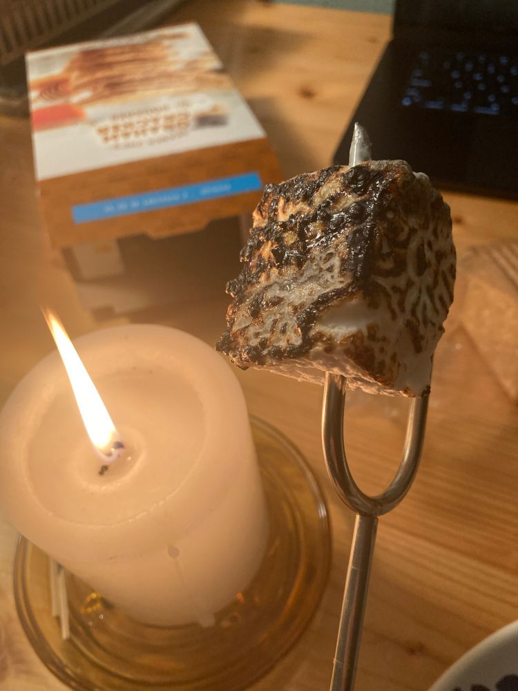 A rectangular homemade marshmallow that’s been lightly charred. Beside it is a candle, and behind is a box of graham crackers. 
