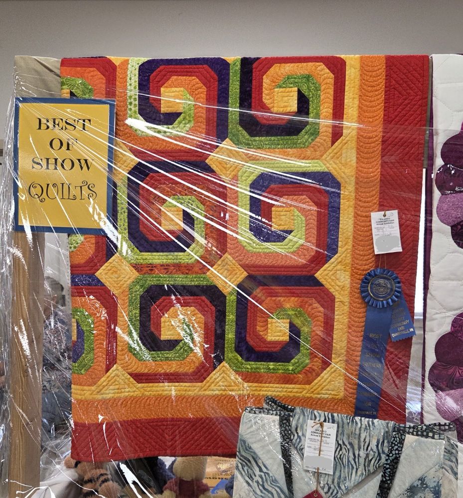 Red, orange, yellow, green, purple spiral quilt with blue ribbon and rosette for Best of Show in Quilts.