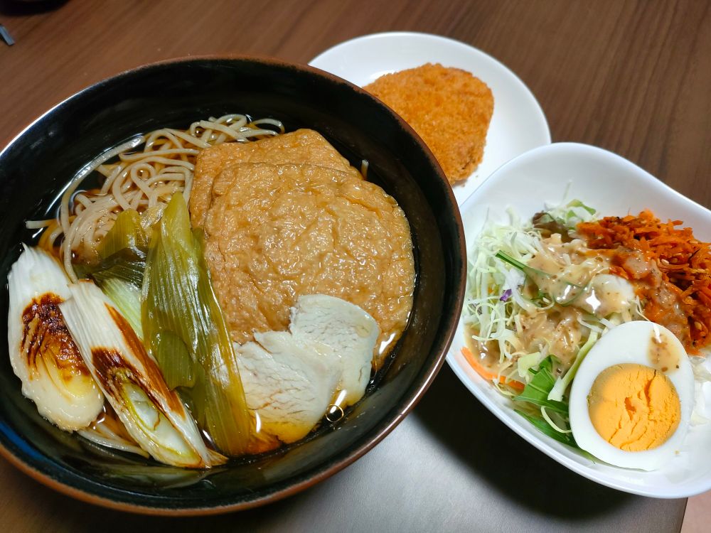 焼きネギと油揚げがのった温かいそば、サラダ、コロッケが写っている。サラダにはゆで卵、千切りキャベツ、人参の千切りが添えられている。
