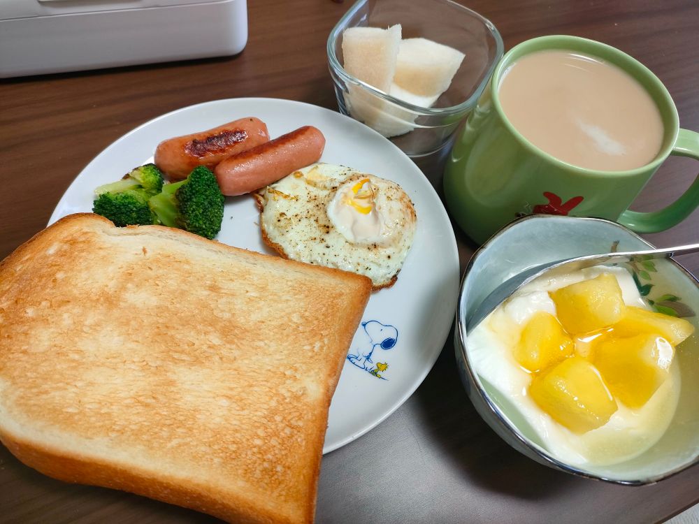 大きなトーストと、目玉焼き、ソーセージ、ブロッコリーが乗ったプレート、パイナップルが乗ったヨーグルト、そしてマグカップに入ったカフェオレが写っている