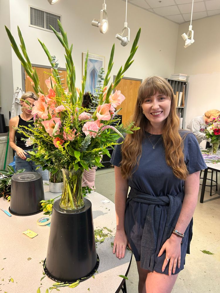 A girl in a blue dress standing next to a tall green and pink bouquet with gladiolas, carnations and eucalyptus with various foliage in a classroom