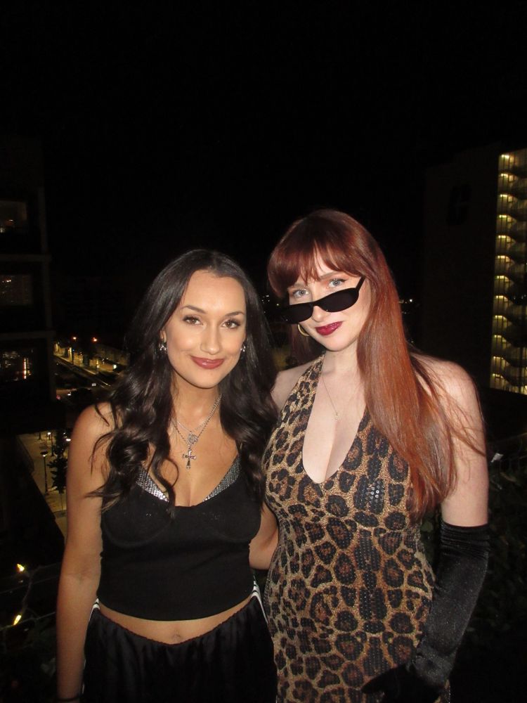 Two girls pose on a balcony in a black tracksuit and cheetah print dress, black gloves and sunglasses