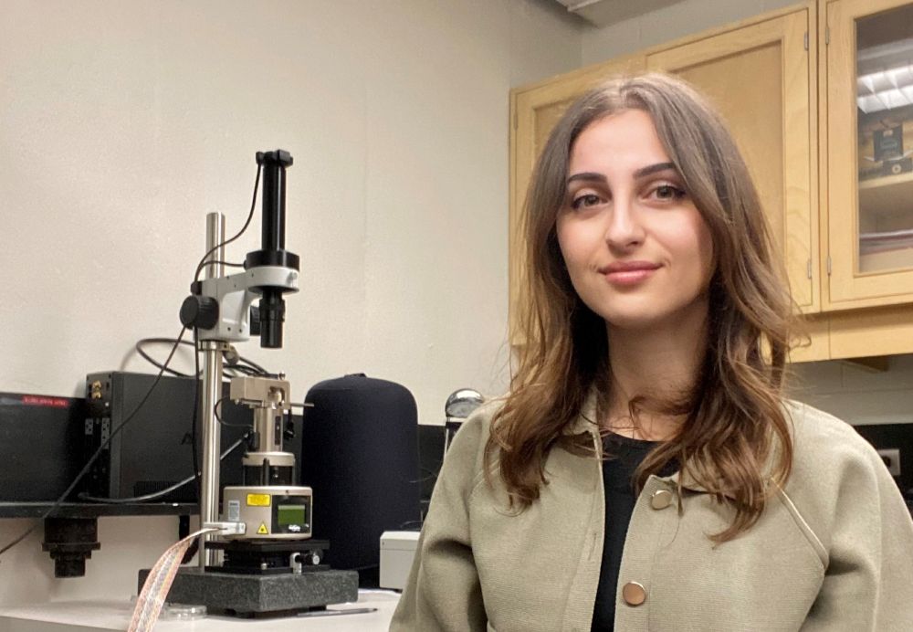 Postdoctoral researcher Alaa Al-Shaer is wearing a beige coat and sitting beside an atomic force microscope