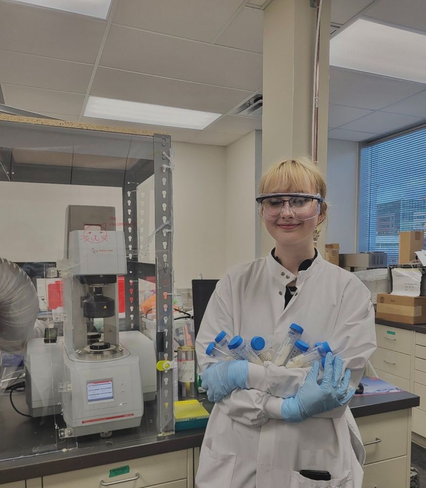 Mariya Konavalova is standing in a science lab wearing a white lab coat, blue nitrile gloves and safety glasses. She's smiling and hugging a bundle of blue-lidded jars of samples.