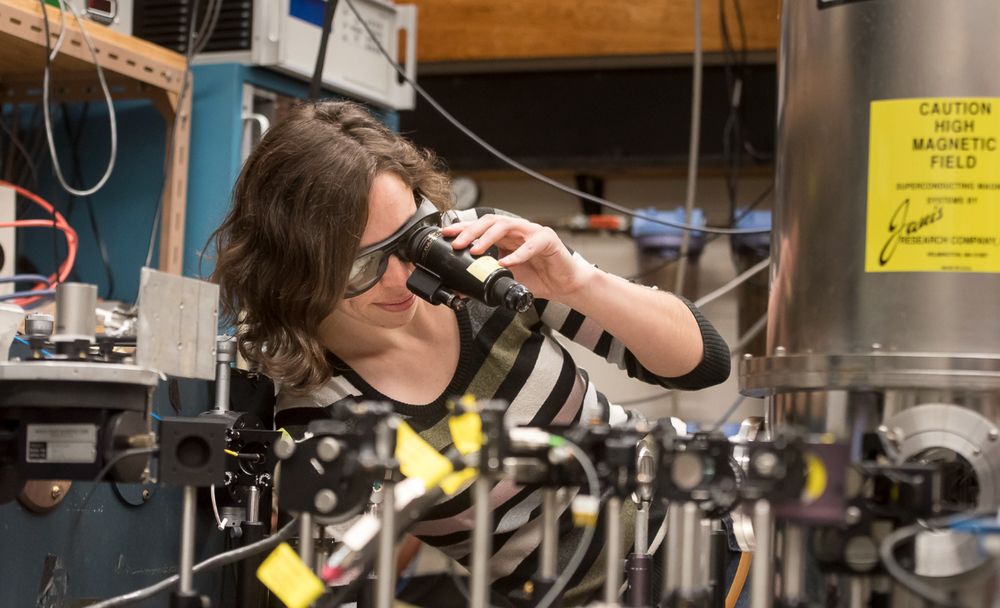 SFU Physics professor Stephanie Simmons in a quantum lab. She is wearing a striped sweater and wearing protective shaded glasses and looking through a special eyepiece at the instruments in front of her.