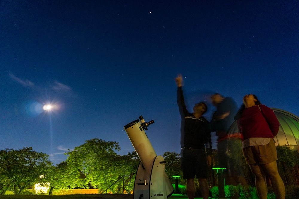 Three people standing beside a telescope and in front of the SFU Trottier Observatory point and look up at the night sky