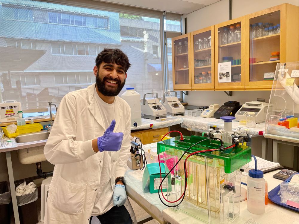 SFU student Arvinder Singh Virk is standing in a science lab, smiling widely. He is wearing a lab coat and purple nitrile gloves and giving a thumbs up sign.