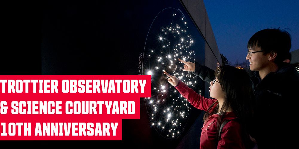 A father and daughter point at an illuminated star chart in the SFU Trottier Observatory Science Courtyard after dark. Their faces are lit by starlight. Overlaid text reads: Trottier Observatory & Science Courtyard 10th Anniversary