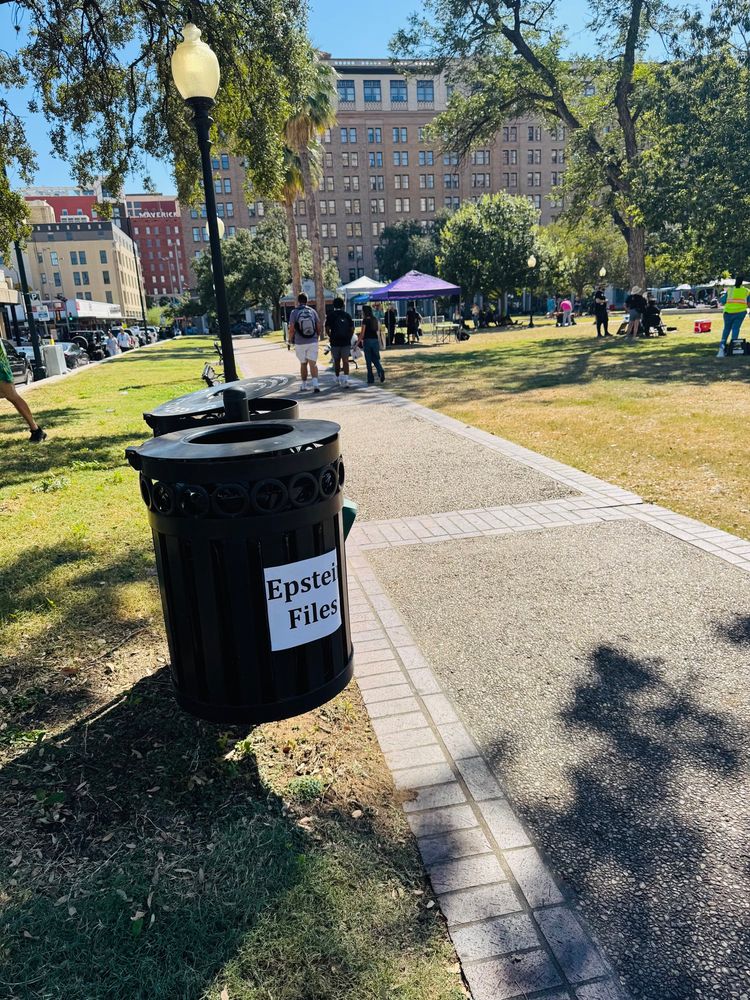 A trash can labeled “Epstein Files” sits along a park walkway, with people and tents visible in the background under sunny weather.