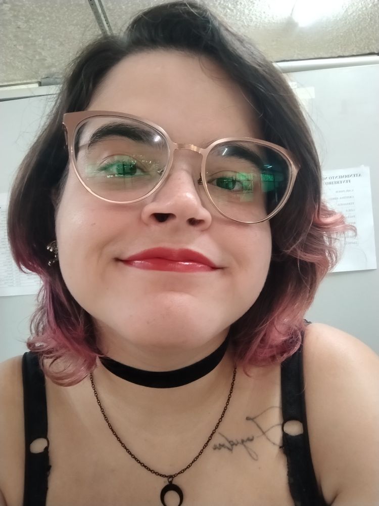 Selfie minha em que estou olhando para a câmera, sorrindo sem mostrar os dentes mas com as bochechas bem elevadas e o queixo levemente erguido. Uso óculos de armação metálica com lentes grandes e levemente gatinho, num tom acobreado. Meu cabelo bate pouco abaixo do queixo e é castanho com as pontas cor de rosa. Uso batom vermelho e uma gargantilha de veludo preta com corrente também preta e um pingente de lua