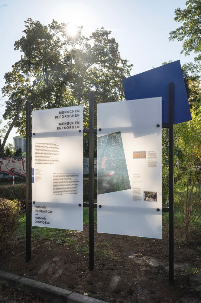 Eine Ausstellungsstation draußen, bestehend aus 2 Glastafeln und einem schräg gestellten blauen Quadrat. Der Titel der Station lautet "Menschen beforschen – Menschen entsorgen".