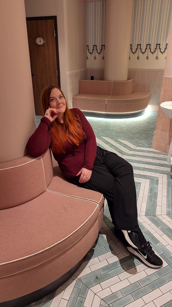 A woman (me) sits on a muted pink rounded bench, wearing a maroon sweater and black pants, sitting with one hand supporting her head leaning back.