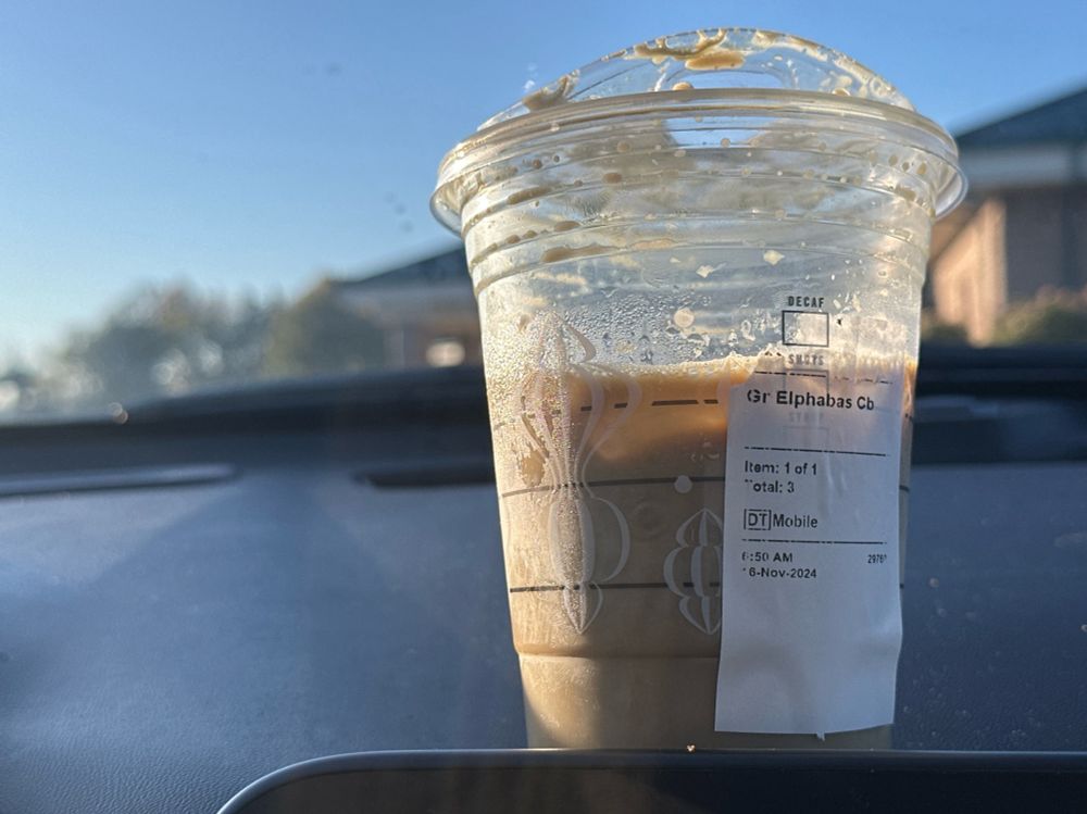 The photo captures a close-up view of a plastic cup of iced coffee sitting on the dashboard of a car. The clear cup has remnants of whipped cream and coffee splatter on the inner surface, with a label adhered to it reading “Grande Elphabas Cold Brew.” The lid is slightly messy, with traces of cream and coffee droplets. The background shows a sunny, clear day, with soft morning light illuminating the scene. A blurred exterior view of trees and a building adds depth to the image. The timestamp on the label indicates the drink was made at 6:50 AM on November 6, 2024.