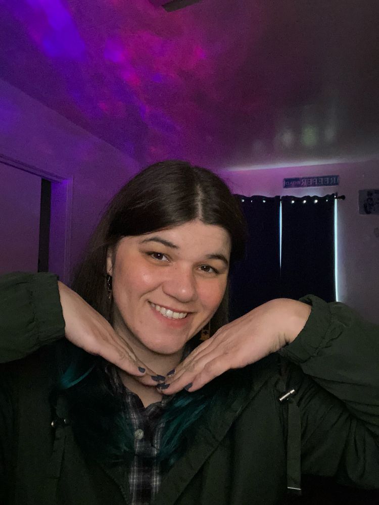 Karma is shown smiling and framing their face with their hands. They are a white person with brown hair dyed blue at the ends that goes right past their shoulders. Karma is wearing wooden guillotine earrings, a black and white flannel and a green rain jacket. Behind Karma there is a bit of light coming through the curtains and the wall has a purplish light shining on it as well.