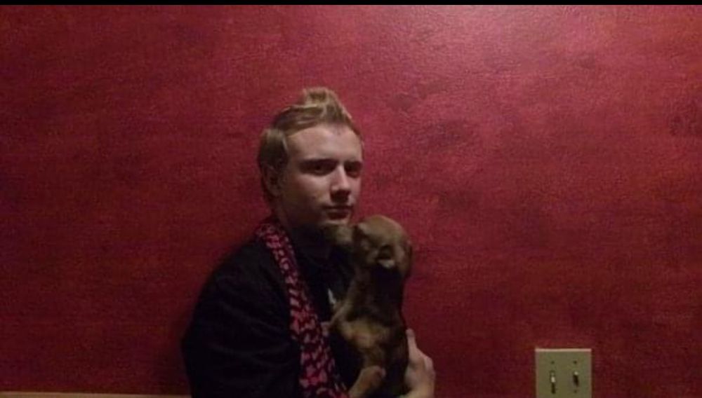 Aaron is shown. He is a white man with short blonde hair. He’s wearing a black jacket and a pink and black zebra print scarf. He’s holding a small brown dog and standing in front of a pink wall.