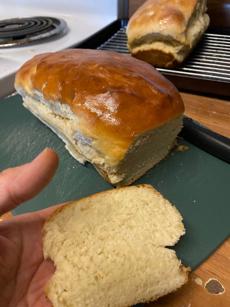One loaf of sandwich bread is on a cutting board with a slice cut off. A white person’s hand is shown holding the slice. In the back a second loaf is sitting on a cooling rack.