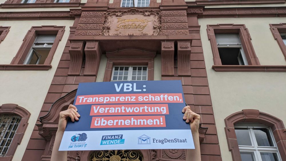 Ein Schild mit der Aufschrift "VBL: Transparenz schaffen, Verantwortung übernehmen!" wird von zwei Händen gehalten. Darüber sieht man das Gebäude des Verwaltungsgerichtshof.