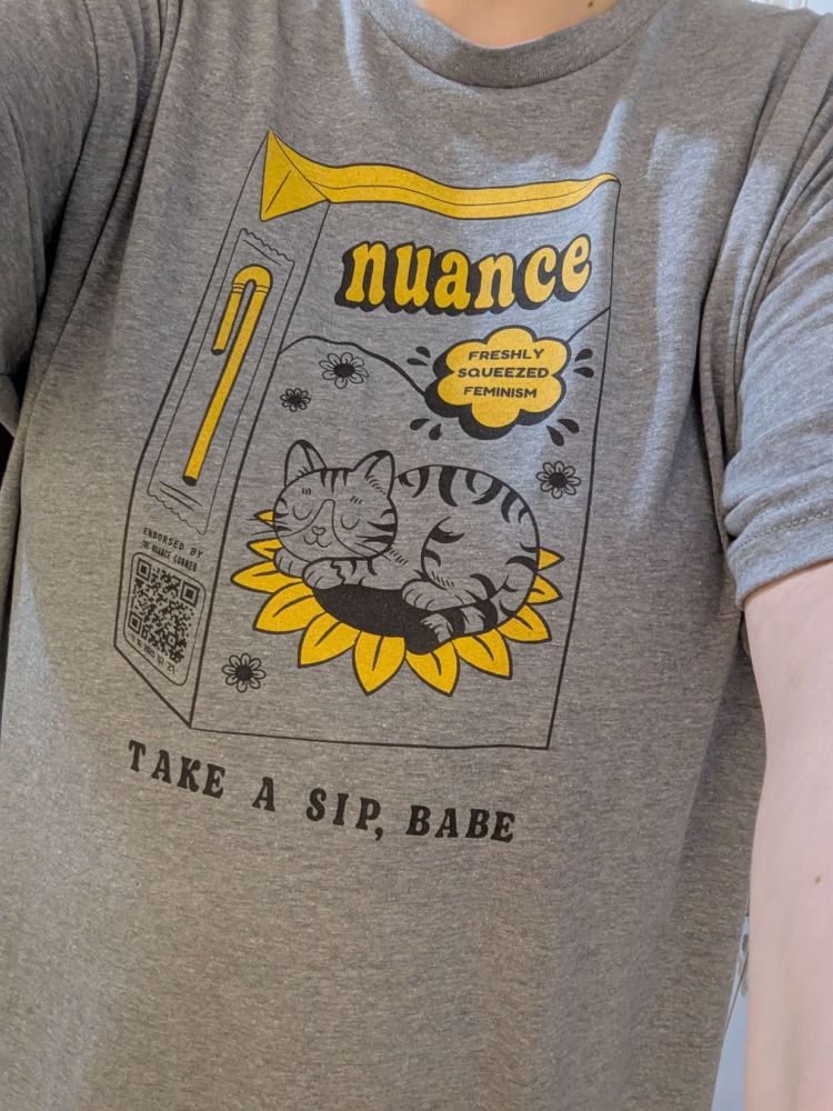 A grey t-shirt with a juicebox design. Logo: Nuance. Body text: Freshly squeezed feminism. Bottom text: Take a sip, babe. A striped cat sits on a sunflower on the front of the juicebox.