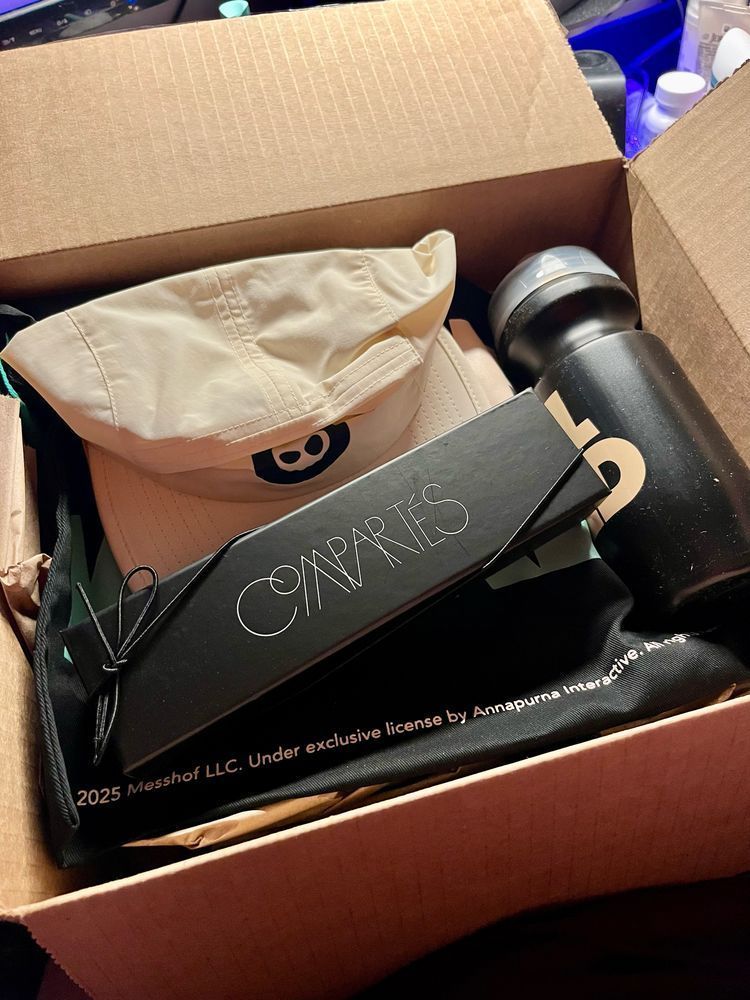 an open cardboard box containing wheel world merch. visible merch includes a plastic water bottle, a hat, a tote bag, and a black box that contains chocolates. 