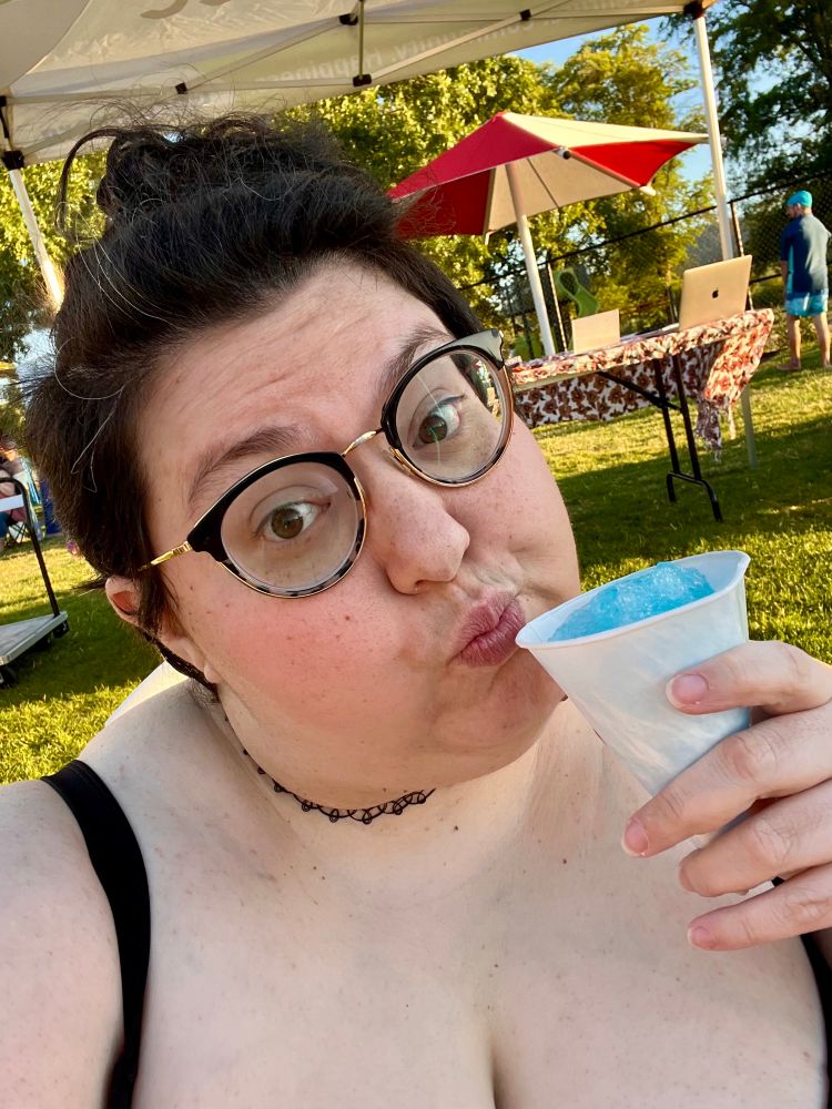a selfie of quinn wearing a simple black bikini top and holding a blue sno cone. their dark hair is piled up on their head, with a visible gray streak at their temple. they wear dark wire frame glasses and make a silly face at the camera. grass is visible behind them. 