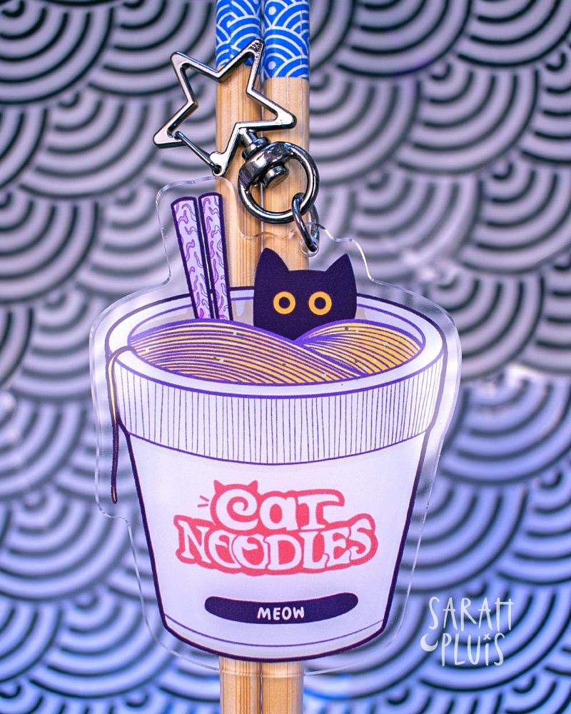 Ein Acrylanhänger mit einem Katzen-Nudel-Motiv liegt auf Essstäbchen.