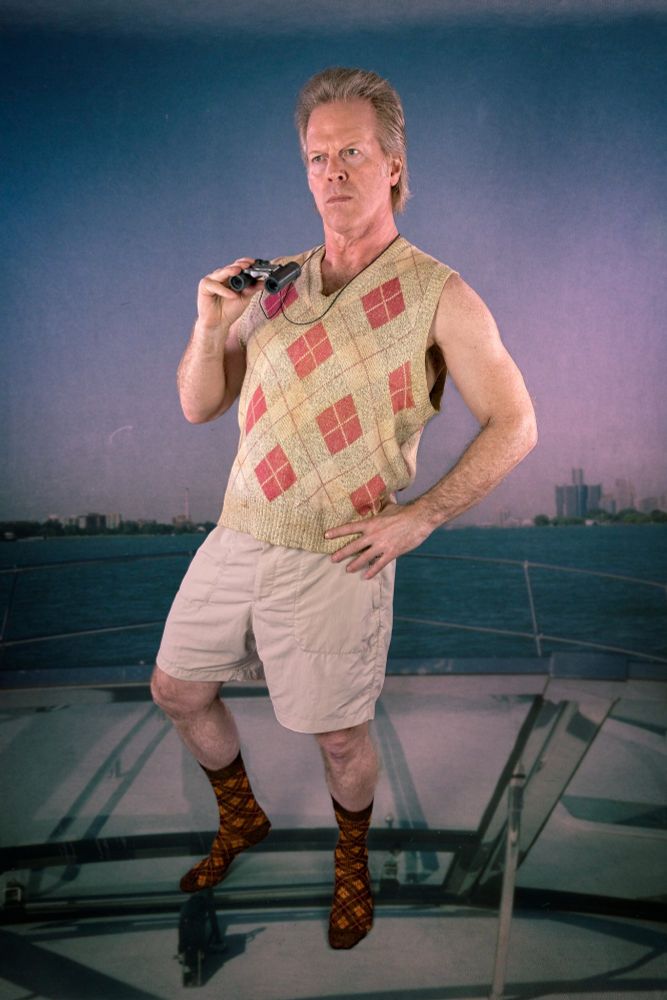 A man wearing an argyle sweater vest and socks from the 1980s and 80s shorts poses with binoculars in hand as he stands on a superimposed background of a boat on a river as 