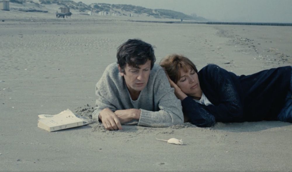 A still from Je t’aime, je t’aime (I Love You, I Love You) by Alain Resnais. On a wide, empty beach under a pale, overcast sky, a man and a woman, Claude and Catrine, lie side by side on the sand. Claude lies on his stomach, propped on his elbows, looking down with a distant, weary expression. Catrine lies close beside him, resting her head against his shoulder. Her eyes are closed. Near Claude’s hand is a small white mouse on the sand; a white paperback lies nearby. In the distance, low dunes, scattered beach huts, and the long shoreline fade into a silent, wintry horizon.