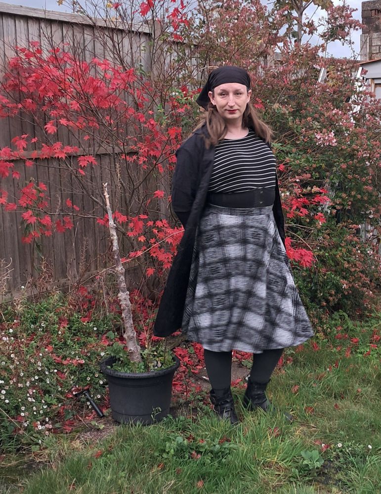 Dae posing in a mostly plack and grey pirate outfit.
Grey leggings and skirt.
Black and white stripped blouse,
Black corset, boots and head scarf.
Black knee-length coat with gold trim.
Standing in front of a bush with red leaves.