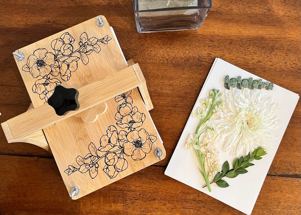 Photo of a flower press next to a paper with a pressed chrysanthemum and two pieces of greenery