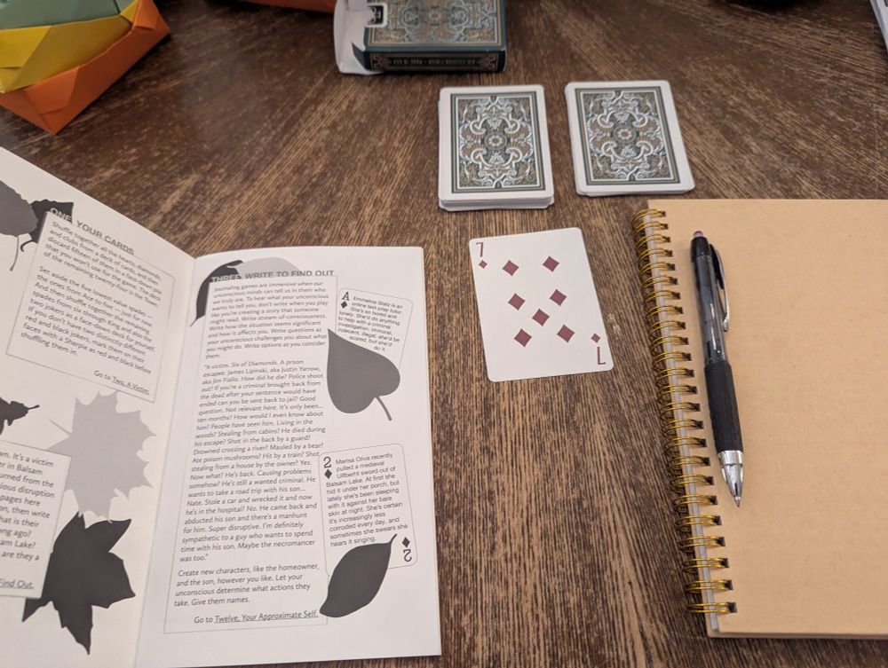 The play materials for a game of the journaling RPG "Balsam Lake Unmurders" laid out upon a table: rule book, playing cards, journal, pen.