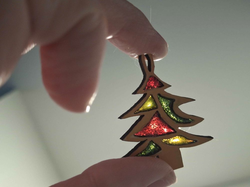 A stylized wooden Christmas tree charm is held up to the light, allowing the red, green, and gold glittery filled cutouts to have a stained-glass effect.
