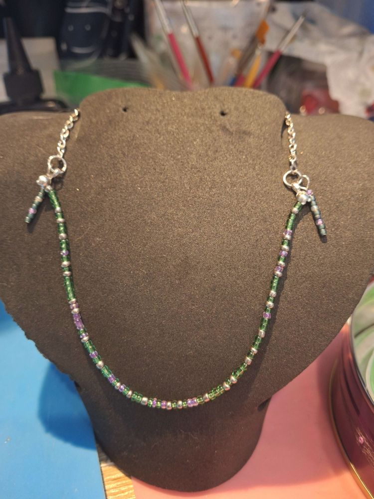 A customizable morse code necklace in green, purple, and white seed beads, with silver chain and two side-dangles with coordinating seed beads. The morse code says 'even the lies especially the lies.' It rests on a black necklace display piece.