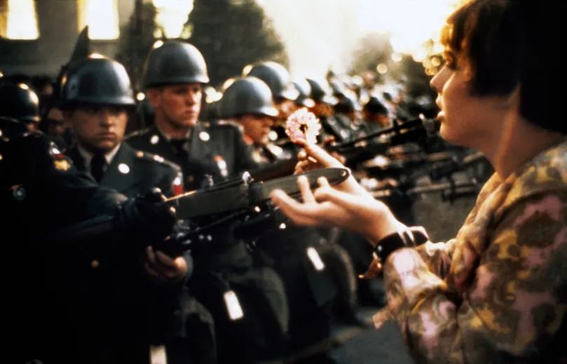 This is an iconic historical photograph, widely known as **"Flower Power,"** taken by photographer Bernie Boston.

The image captures a tense yet poignant moment during an anti-Vietnam War protest at the Pentagon on October 21, 1967.

In the frame, a young protester with short dark hair is seen on the right, extending her hands toward a line of soldiers. She is gently placing a single carnation into the barrel of a rifle held by one of the soldiers.

Facing her is a formidable line of military police or National Guardsmen in full uniform, equipped with steel helmets and holding rifles with fixed bayonets, forming a human barricade. The soldiers appear very young and have stoic, serious expressions.

The photograph is renowned for its powerful symbolism and stark visual contrast:

* **Peace vs. War:** The soft, fragile flower against the hard, menacing steel of the rifle.
* **Non-violence vs. Force:** The gentle, peaceful gesture of the protester in the face of armed authority.
* **Humanity vs. Institution:** The individual's act of connection towards the uniformed, anonymous line of soldiers.

The image has become a defining symbol of the 1960s counterculture movement and the philosophy of non-violent resistance, powerfully encapsulating the clash between the peace movement and military force.