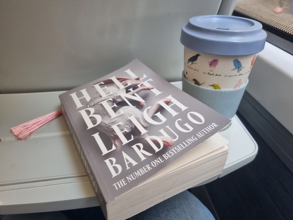 A picture of a copy of Hell Bent by Leigh Bardugo and a keep cup of coffee on a train tray table.