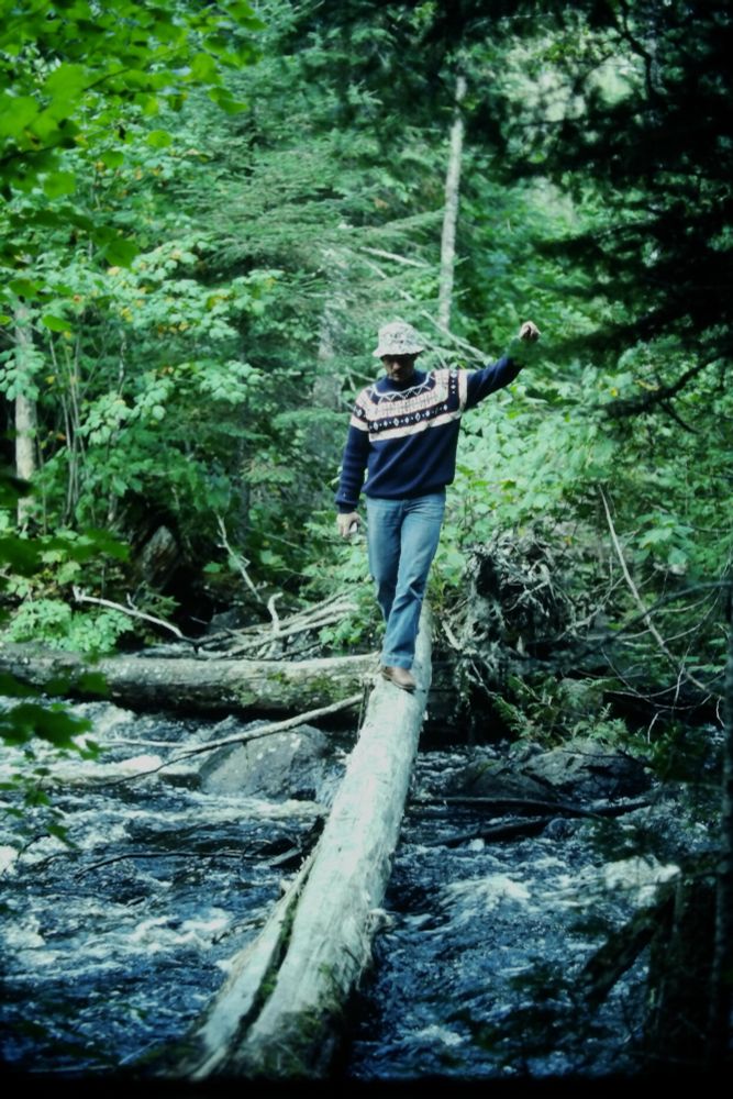Ned Johnston traversing a fallen log crossing a river.