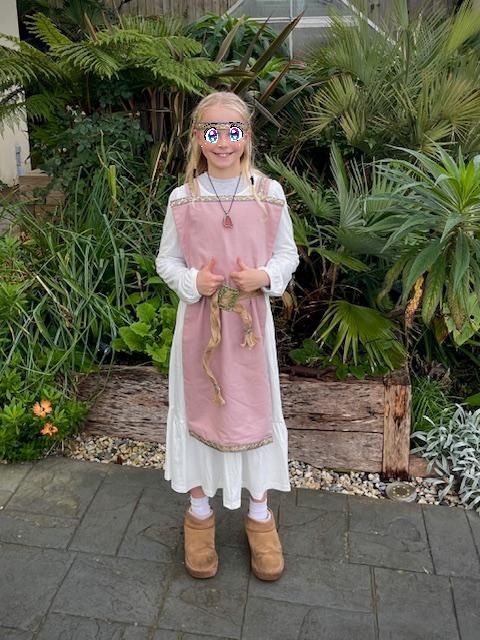 A photograph of smiling Little Wiggle in her Anglo Saxon outfit. She has a white dress as a linen underdress, and a dusky pink (lambs wool" tabard, trimmed with blue and gold braid at the top, bottom and shoulder straps. Her belt is made with jute ribbon plaited together, the ends finished with more braid, and a gold buckle. She is wearing a large amber and silver pendant, and Ugg boots.