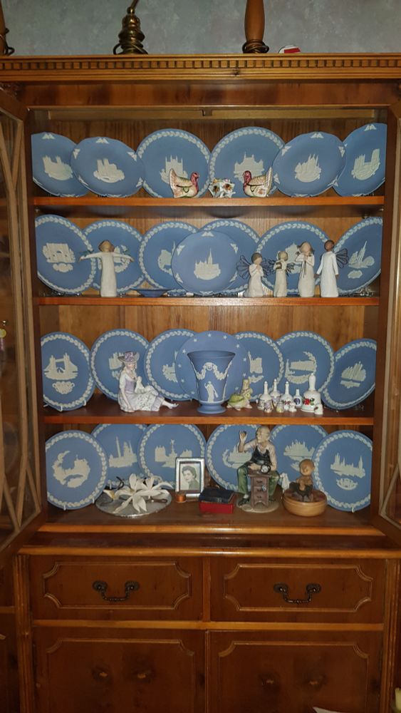 A photograph of an open large wooden dresser unit, showing a large collection of blue and white Wedgwood plates, amidst other ornaments of things like a porcelain clockmaker, Willow Tree angels and two ceramic turkeys that are actually salt and pepper shakers.
