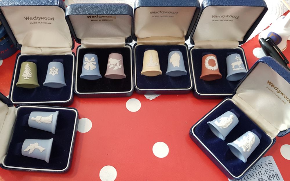 A photograph of 12 boxed Wedgwood coloured jasperware Christmas thimbles, in various colours, in pairs. Blue, green, pink, yellow and terracotta form the base colour, and each has a raised white motif of various Christmas themes, including a tree, snowflake, robin, Santa, angel, shepherds and Mary.