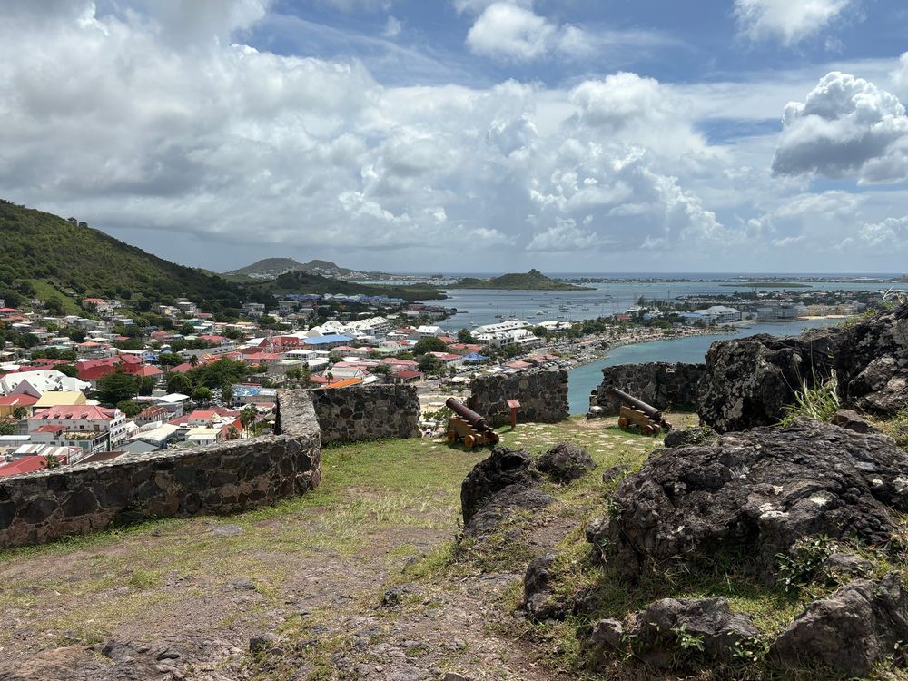 Low stone walls and two cannon overlook brightly colored roofs and blue water below. There are some black rocks in the foreground, probably ruins of a former building.