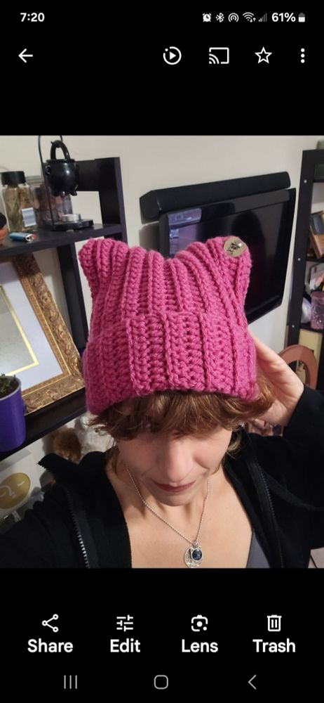 A person stands in a fashionable pose, head down, hand behind their head, a shelf in the background, showing off a pink cat hat with a logo tag on the ear. The hat a ribbed crochet, with a fold up brim is of a raspberry pink shade.