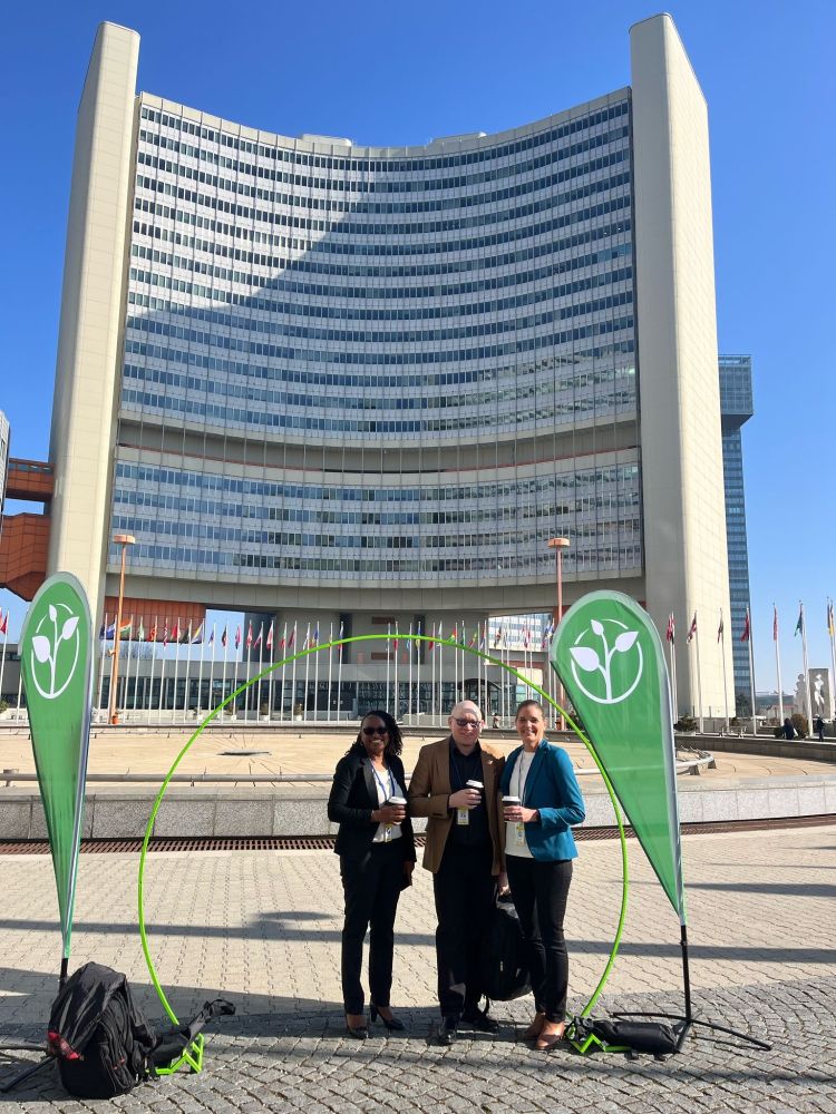 Two women and a man stand in front of a large, glass fronted building- the UN Offices in Vienna - in between two banners featuring Zero Project conference logos