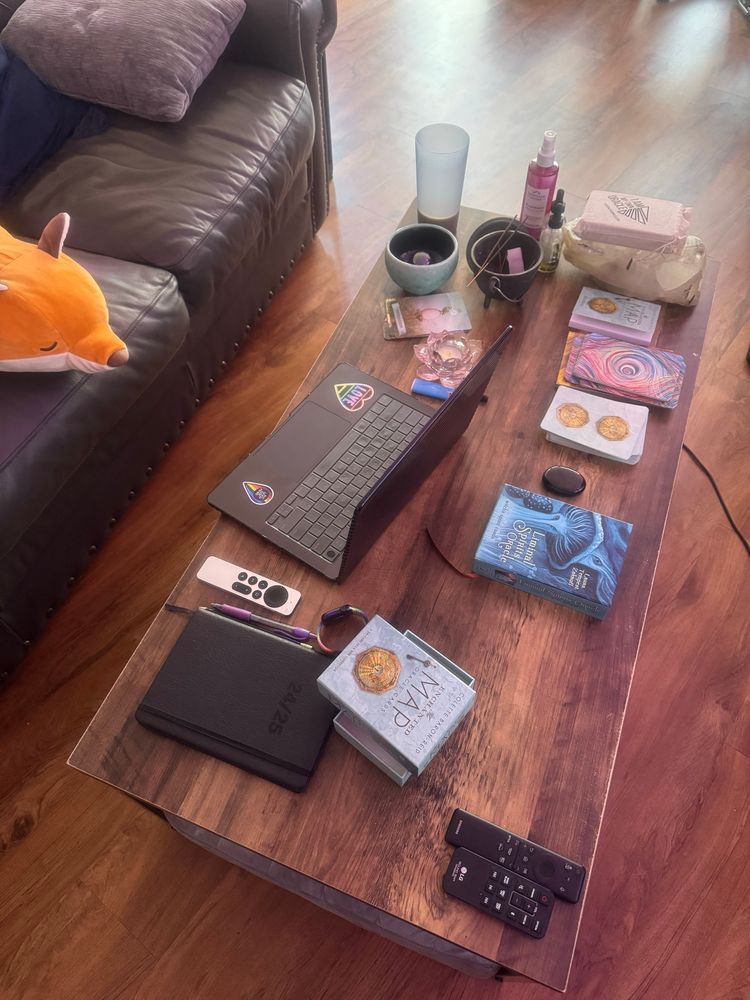 Coffee table workstation. Laptop, journal, and occult curios including gemstones, candles, rose water, and oracle cards.
