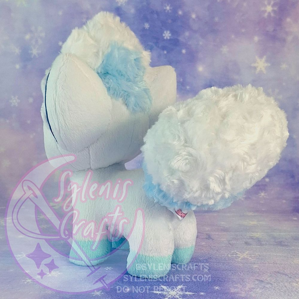 A chibified Alolan Vulpix Plush made with minky fabric. It is standing facing away from the camera to better show the tails and the back of its head. It stands on a purple backdrop patterned with snowflakes. 
