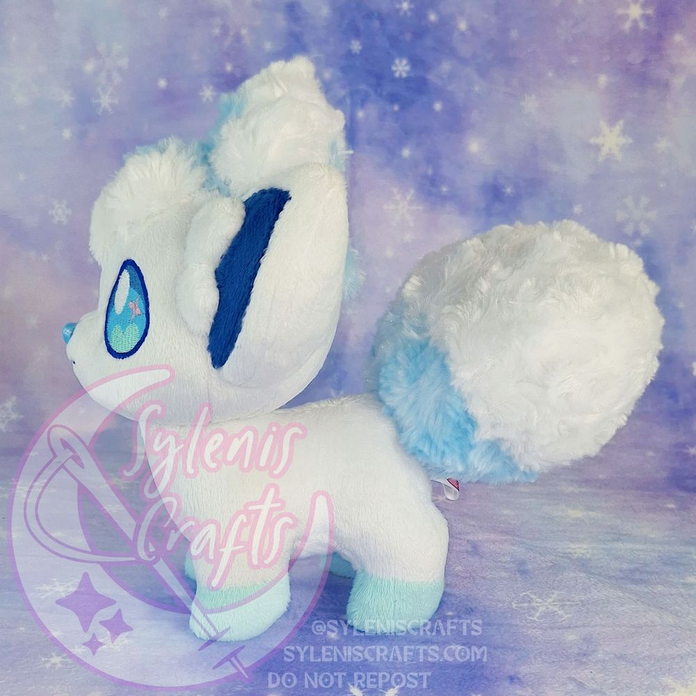 A chibified Alolan Vulpix Plush made with minky fabric. It is standing facing left, its head is slightly up tilted to mimic the official artwork. It stands on a purple backdrop patterned with snowflakes. 