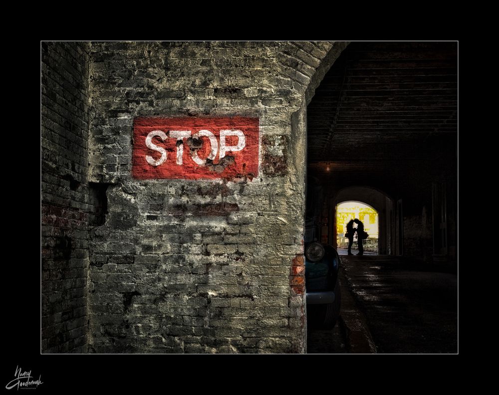 We're at Alcatraz Prison with a brick wall printed with a rectangular red STOP sign on the left, and dark archway with lit arch in background with couple standing and touching heads in silhouette on the right. "Arch" is the theme for #BlueSkyArtShow on March 29, 2025. Copyright Nancy Goodenough 2012