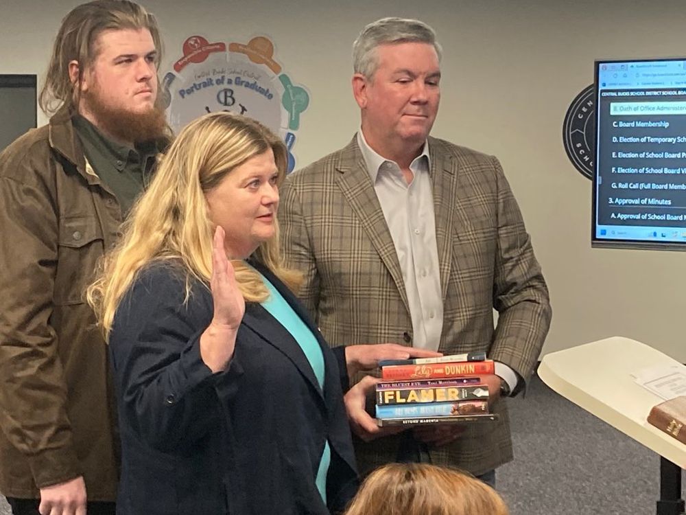 Karen Smith being sworn into her new position. She has her right hand in the air and left hand on a stack of books that have been banned in her area. Two men stand to her left, one holding the stack of banned books.