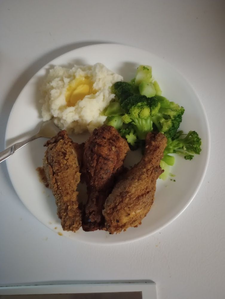 A photograph of a meal on a plate: Three fried chicken legs done golden brown at the bottem of the picture,with steamed broccoli to the upper right, and mashed potatoes with a melting pat of butter to the upper left. The background, and the plate, is white, and a fork protrue from the mashed potatoes.