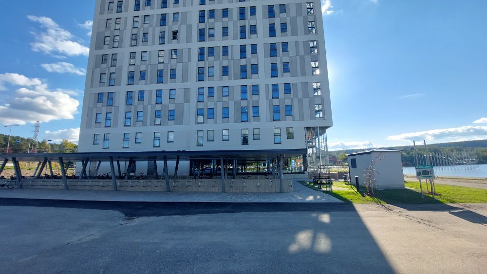 Korkea, iso, harmaa kerrostalo. Etualalla iso pyöräkatos, jossa jykeviä runkolukitustelineitä.